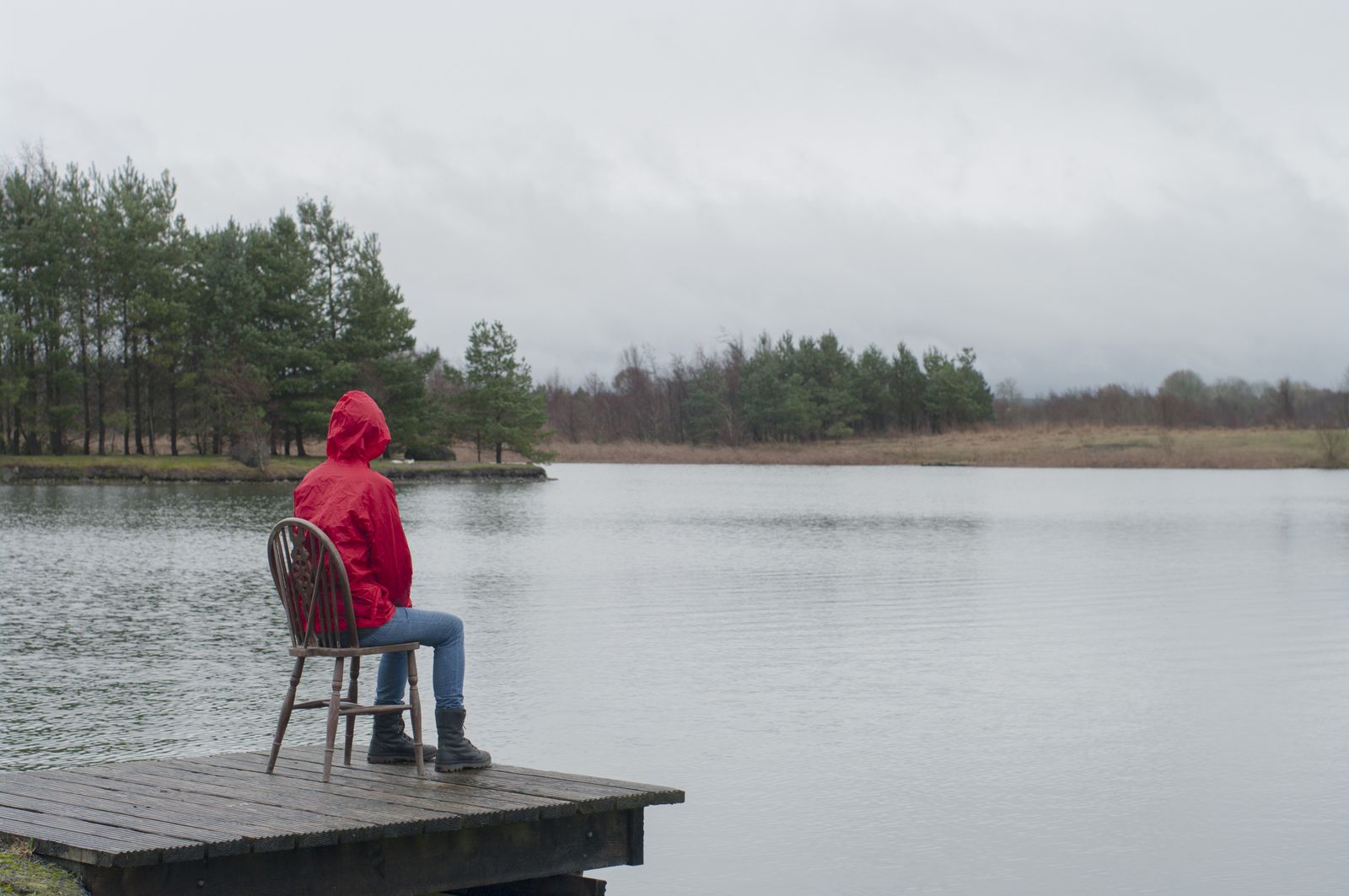 A figure in a red raincoat sits on a wooden chair at the end of a jetty, facing a lough